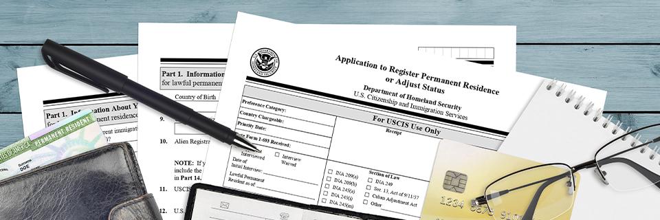 A Form I-485 Application to Register Permanent Residence or Adjust Status, with glasses, a pen, a U.S. Green Card, a wallet, and other items on a desk.