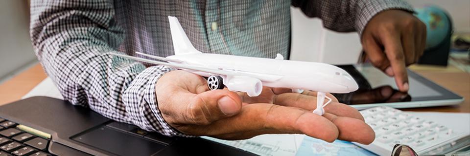 A hand holding a model airplane above a desk with a tablet and a computer, symbolizing moving to the U.S. and getting a permanent residence green card.