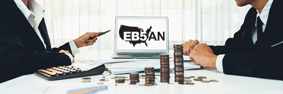 Two EB-5 experts sitting at a desk with stacks of coins and a computer between them, with the EB5AN logo showing on the computer screen.