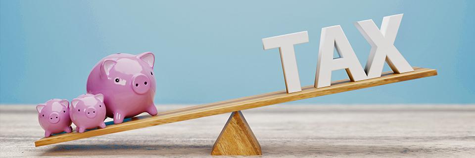 A wooden balance with piggybanks on one side and the letters TAX on the other.