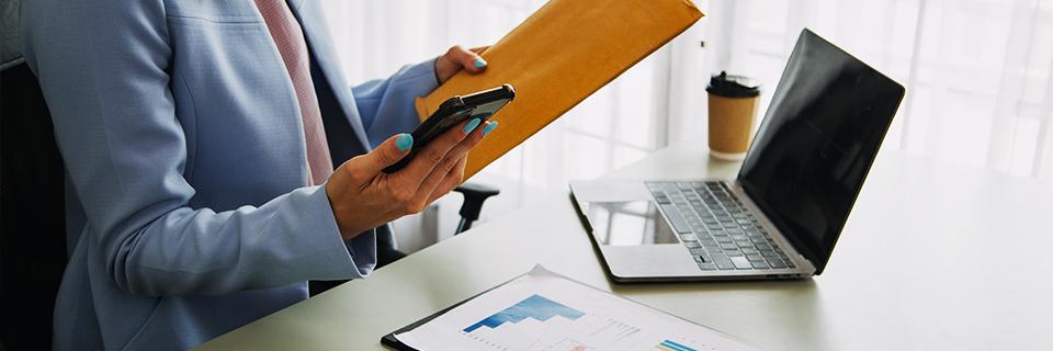 An investor at an office with a laptop and an envelope, looking at her phone, researching EB5 step by step process.