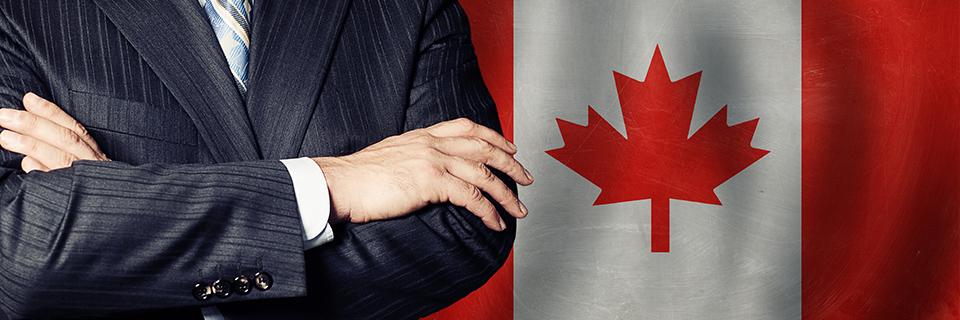 A person in a suit with their arms crossed in front of a Canadian flag.