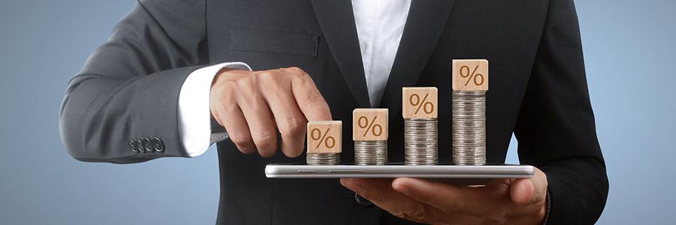 A hand holding a tablet with stacked coins and blocks with a percentage signs on them.