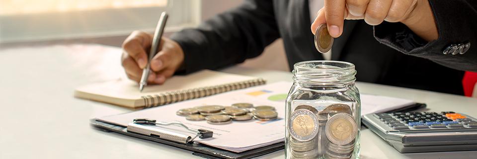 A person putting coins into a jar while making calculations on a notebook, symbolizing economic development through the regional center program.