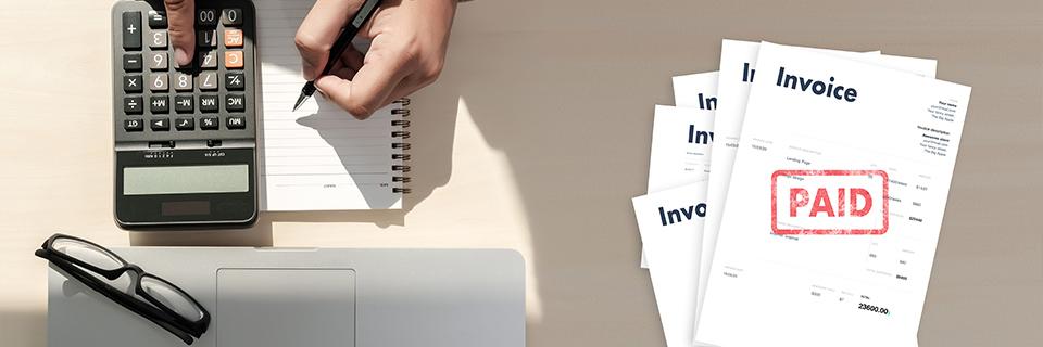 A person writing on a notebook and using a calculator, with paid invoices on the side.