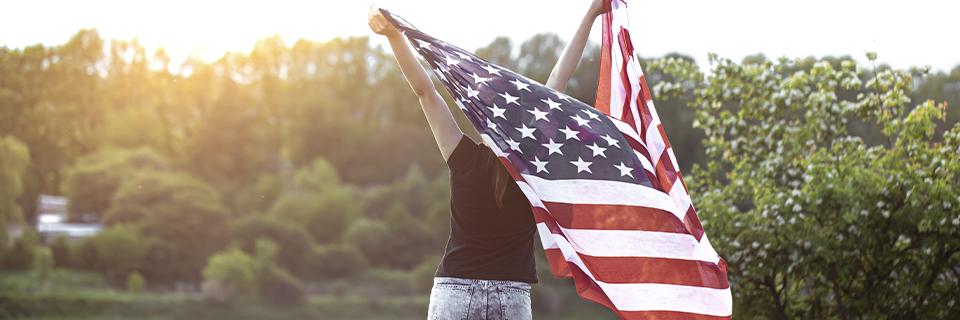 A green card holder standing outdoors with her hands in the air, holding a U.S. flag.