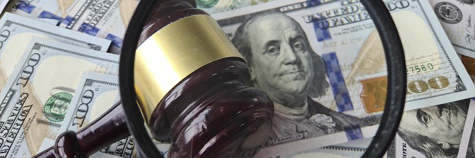 A close up of a magnifying glass and a judge's gavel on a stack of U.S. dollars.