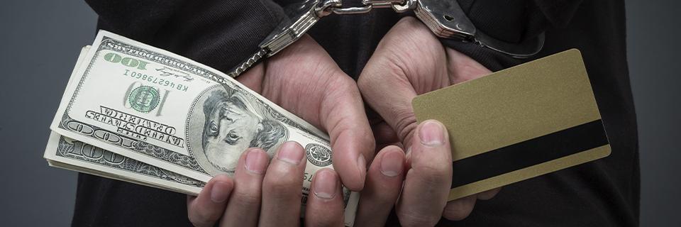 A close-up of hands holding dollars and a credit card in handcuffs, symbolizing fraud or money laundering.