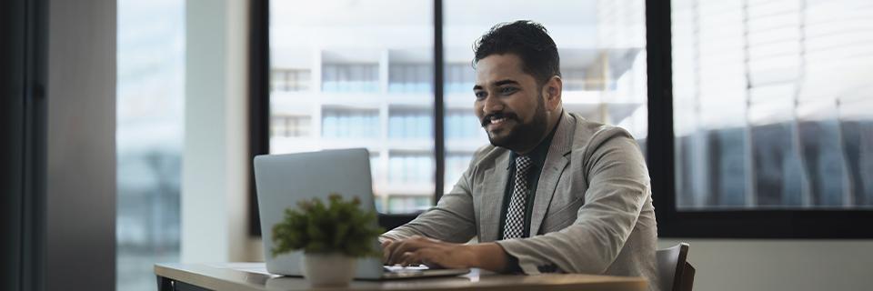 A smiling Indian businessman working on a laptop in an office, symbolizing professional growth and opportunities through U.S. immigration programs like EB5.