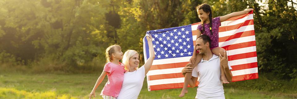 A family celebrating in a scenic park, holding a US flag. The EB5 program provides a pathway to U.S. permanent residency, offering families the chance to build a future together in America.