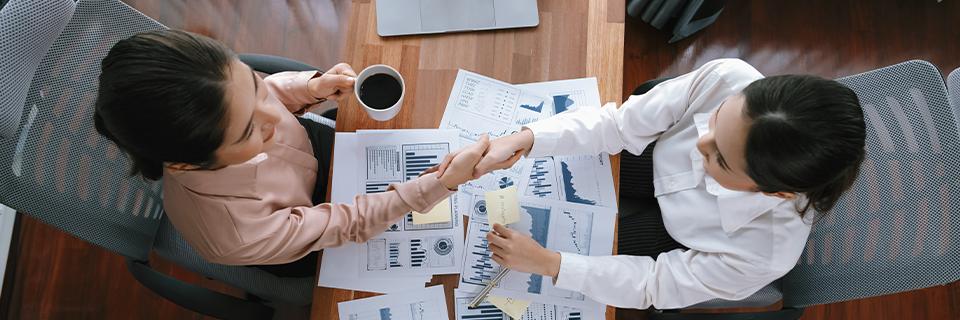 Two businesswomen shaking hands over financial documents and charts, showcasing partnership and collaboration in EB5 projects.