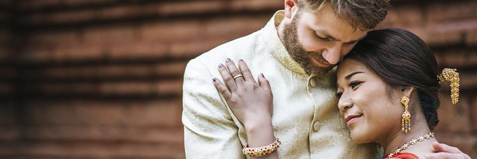 A multicultural couple in traditional wedding attire embraces, symbolizing international marriage and the process of obtaining a U.S. Green Card through a spouse.