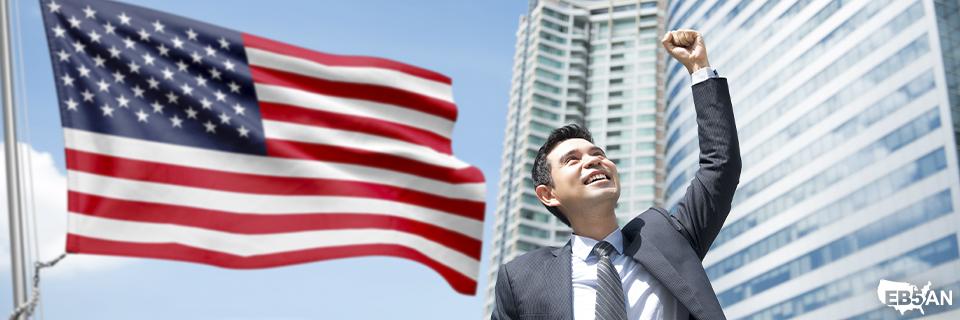 A smiling Singaporean businessman raises his fist in celebration, with the U.S. flag waving in the background. Skyscrapers symbolize career and investment opportunities in the United States. EB5AN logo in the corner.