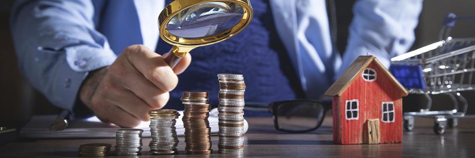 Businessperson analyzing stacked coins with a magnifying glass next to a small model house, illustrating EB5 investment capital and source-of-funds documentation.