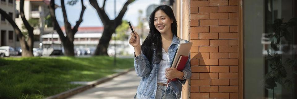 International student smiling outside a school building, holding books and a pen, representing F1 visa holders pursuing education in the U.S. on a non-immigrant visa.