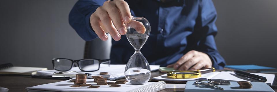 A businessman flips an hourglass next to stacked coins and documents, symbolizing time-sensitive EB5 investment decisions and financial planning.