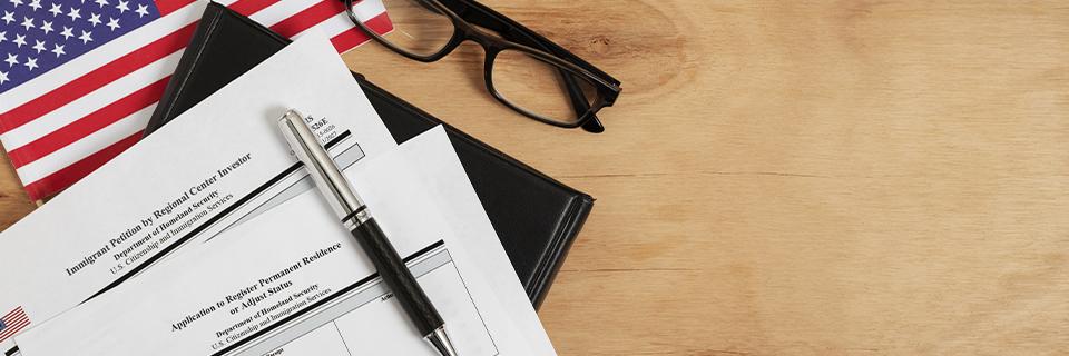 EB5 immigration forms including Form I-526E and Form I-485, placed beside a pen and U.S. flag, symbolizing concurrent filing for Green Card applicants.