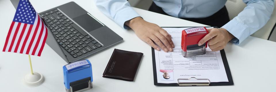 An immigration officer stamping “REJECTED” on a visa application form beside an “APPROVED” stamp, a U.S. flag, and a laptop—symbolizing the contrasting outcomes of EB5 petition processing.