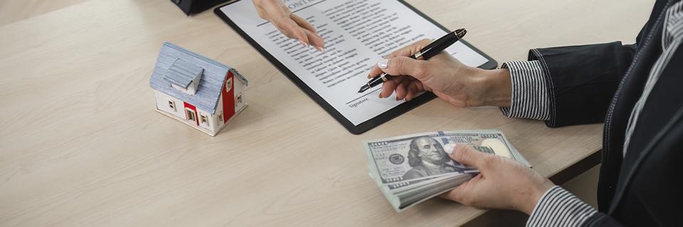 Close-up of a real estate transaction involving a mini house model, contract signing, and a stack of hundred-dollar bills, representing EB5 capital structure and third-party loan involvement.