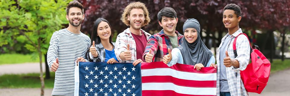 Smiling international students holding American flag and giving thumbs up, representing success through the EB5 visa path to U.S. permanent residency.