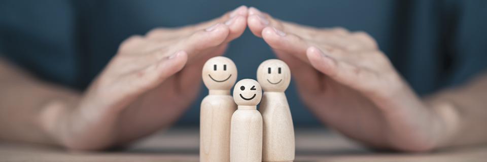 Hands gently shielding a smiling wooden family figure set, illustrating the EB5 visa’s family benefits, including Green Cards for spouses and children.