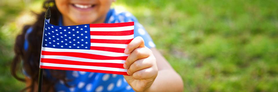 A smiling child holding a small American flag outdoors, symbolizing the dream of U.S. citizenship and a future in the United States.
