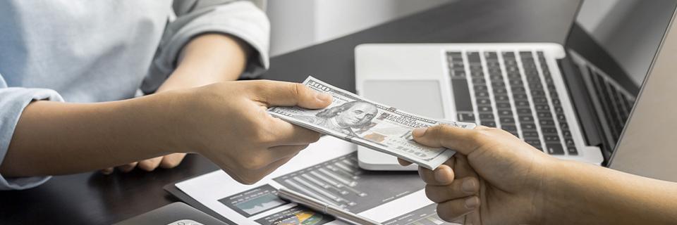 Close-up of cash being handed over at a business desk with financial charts and laptop, illustrating EB5 loan repayment and guarantor obligations.