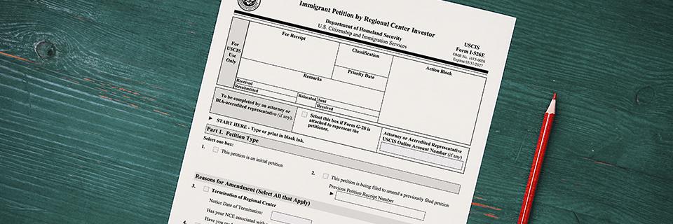 USCIS Form I-526E petition for EB5 regional center investors placed on a wooden desk with a red pencil, highlighting key immigration paperwork.