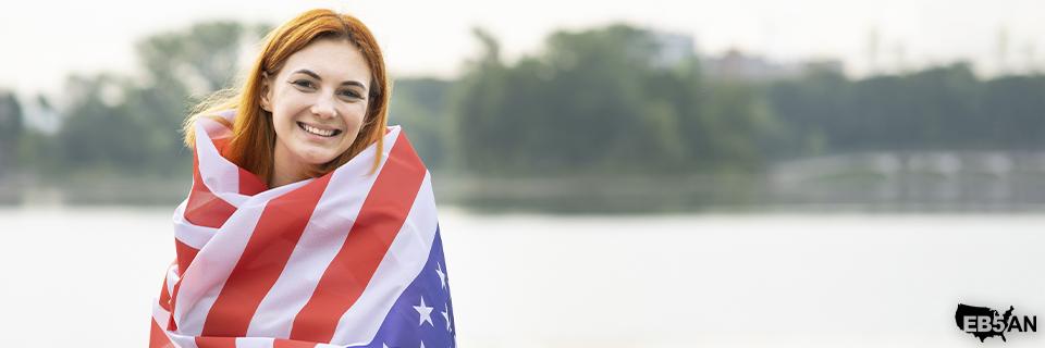 A smiling woman wrapped in an American flag, representing pride in U.S. citizenship and belonging to the American community.