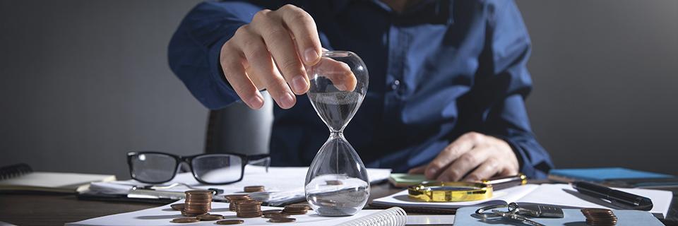 A person measuring time with an hourglass beside coins and paperwork, symbolizing EB5 investors waiting for project repayment and financial returns.
