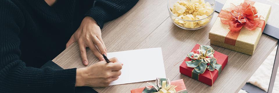 Person writing a letter at a table with wrapped gifts nearby, illustrating preparation of a notarized gift letter required for EB5 source of funds documentation.
