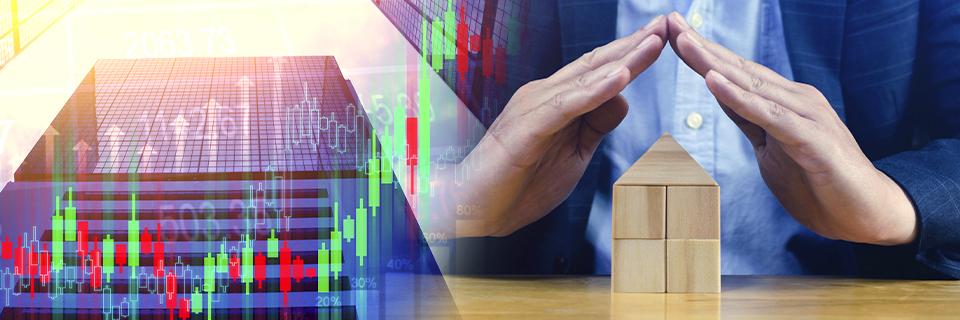 Hands forming a protective shape over wooden house blocks with financial charts in the background, representing EB5 investors using U.S. investment to hedge against inflation and economic instability.