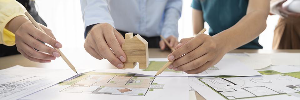 Hands placing a small wooden house model over detailed floor plans, illustrating EB5 project planning, real estate development strategy, and long-term housing demand supporting job creation.